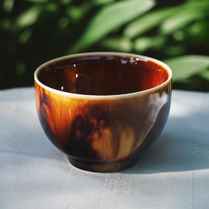 Puede incluir: Un cuenco de cerámica con un esmalte marrón brillante. El cuenco tiene una forma redondeada con un borde ligeramente ensanchado y una base oscura. El interior es de un marrón oscuro intenso y el exterior tiene un efecto veteado.