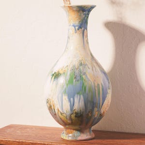 May include: A ceramic vase with a bulbous body and a flared rim, featuring a multi-colored drip glaze in shades of blue, green, yellow, and white. A small bunch of dried, thin-stemmed flowers with tiny white blossoms is placed inside the vase.