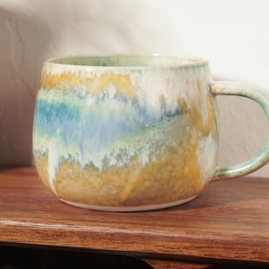 May include: A ceramic mug with a rounded body and a side handle, featuring a reactive glaze finish in blended shades of ochre yellow, teal blue, and creamy white, with the colors layered in a horizontal, flowing, and mottled pattern.