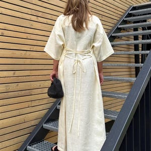 May include: A long, cream-colored dress with short sleeves and a tie-back waist. The dress is worn by a person ascending metal stairs. A small black handbag is held in the left hand. The background features wooden siding.