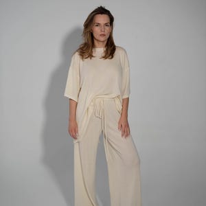 May include: A woman is wearing a cream-colored, loose-fitting, knit top and matching pants. The top has a round neckline and short sleeves. The pants have a drawstring waist and wide legs.