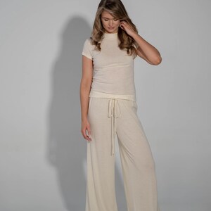 May include: A woman is wearing a cream-colored, short-sleeved top and matching wide-leg pants. The pants have a drawstring waist.