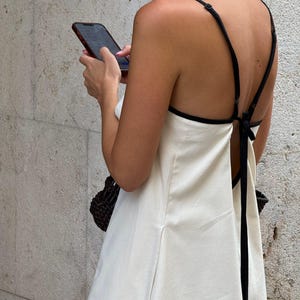 以下が含まれることがあります： 黒いトリムとスパゲッティストラップが付いたクリーム色のドレス。ドレスは背中が大きく開き、裾の近くに小さな円形のロゴが付いています。黒いハンドバッグが背景に見えます。