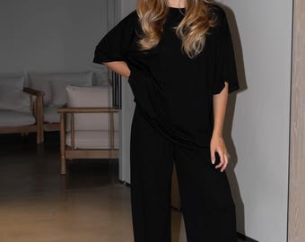 May include: A woman wearing a black, loose-fitting, long-sleeved top and matching wide-leg pants. She is standing in a room with light wood floors and white walls.