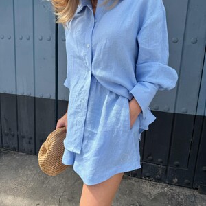 May include: A pale blue linen shirt and shorts co-ord. The long-sleeved shirt is buttoned and the shorts have pockets. A straw hat is held in the hand. The outfit is worn in front of a blue door.