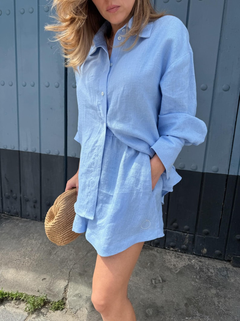 May include: A pale blue linen shirt and shorts co-ord. The long-sleeved shirt is buttoned and the shorts have pockets. A straw hat is held in the hand. The outfit is worn in front of a blue door.