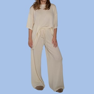 May include: A woman is wearing a cream-colored, loose-fitting, short-sleeved top and matching wide-leg pants. The pants have a drawstring waist.