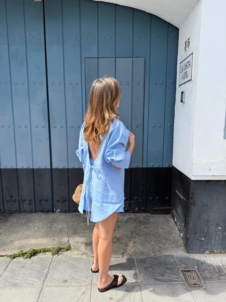 May include: A light blue linen dress with an open back and tie detail. The dress has long sleeves and is paired with black sandals and a straw bag. The background features a blue door and a white building with the text "LA CASITA AZUL".