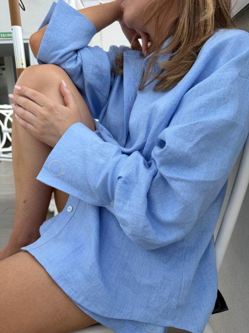 May include: A pale blue linen shirt with long sleeves and button fastenings. The shirt has a loose fit and is made of a lightweight material. The sleeves are cuffed at the wrists. The top buttons are undone.