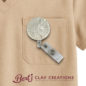May include: A sand dollar badge reel with a white strap and a metal clip is attached to a tan pocket. The badge reel is a polymer clay ornament. The text "Bert's CLAY CREATIONS" is at the bottom of the image.