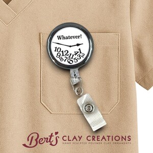 May include: A round, black and white badge reel with the text "Whatever!" and a clock face design. The badge reel is attached to a white strap with a snap closure and is clipped to a tan pocket.