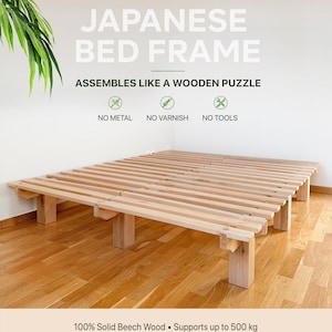 Op de afbeelding: Een lichtgekleurd bedframe van massief beukenhout met een lattenbodem. De tekst luidt: JAPANESE BED FRAME, ASSEMBLES LIKE A WOODEN PUZZLE, NO METAL, NO VARNISH, NO TOOLS, 100% Solid Beech Wood, Supports up to 500 kg. Het frame staat laag.