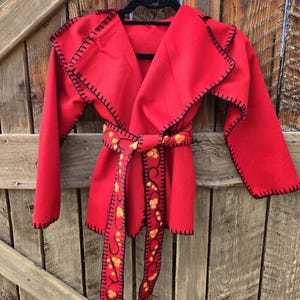 May include: A red wrap coat with black stitching and a matching belt. The belt features a decorative pattern with yellow and orange accents. The coat is hanging on a black hanger against a wooden background.