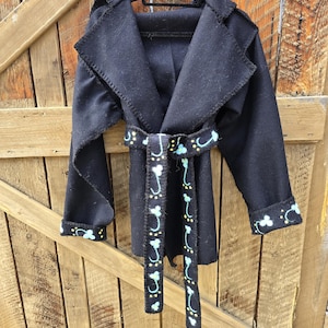 以下が含まれることがあります： ショールカラーとマッチするベルトが付いた黒いコート。ベルトと袖口には、水色と黄色の花の繰り返し模様が施されています。コートは、木製のドアを背景に、黒いハンガーに掛けられています。