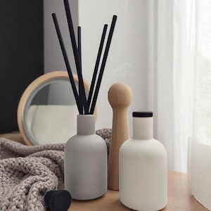May include: Aesthetic home decor featuring a gray diffuser bottle with black reeds, a white bottle with a black top, and a wooden decorative piece. The items are arranged on a wooden surface with a knitted throw and a round mirror in the background.