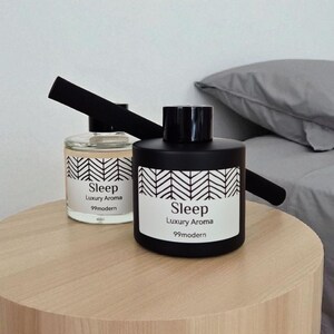 May include: Two aroma diffusers labeled "Sleep Luxury Aroma 99modern" sit on a wooden side table. One is clear with a black top, the other is black. Both have a white label with black text and a chevron design.