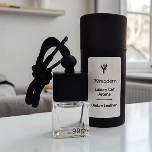 May include: A clear, square car air freshener with a black cap and a black cord, next to a black cylindrical container. The air freshener has the text "99m" on the front. The container has a white label with the text "99modern Luxury Car Aroma Ombre Leather".