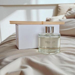 May include: A clear glass diffuser bottle with a silver cap and the text "99modern" and "Seville" printed on the front. A white box and a bundle of wooden reeds sit behind the bottle. The items are on a beige surface.