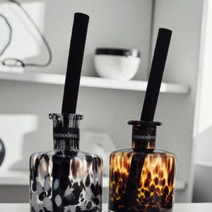 Op de afbeelding: Twee decoratieve glazen flessen met zwarte rietjes. Eén fles heeft een zwart-wit stippenpatroon, de andere een bruin en amberkleurig stippenpatroon. De tekst "99modern" is op de voorkant van elke fles gedrukt.