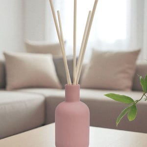 May include: A pink essential oil diffuser with several light-colored reeds. The diffuser sits on a wooden table, with a neutral-toned sofa and pillows in the background. A few green leaves are visible on the right.