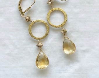 May include: Gold earrings with a textured circle and a teardrop-shaped topaz gemstone. The earrings have a leverback closure.