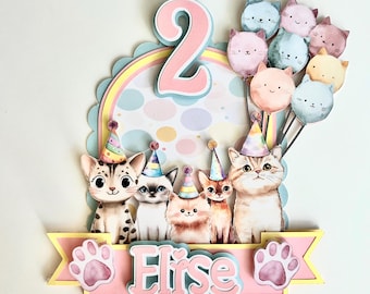 Può includere: Decorazione colorata per torta di compleanno con gatti dei cartoni animati con cappelli da festa. La decorazione include il numero "2", palloncini a forma di gatto e il nome "Elise" su uno striscione, con impronte di zampe. Il design ha una combinazione di colori pastello.