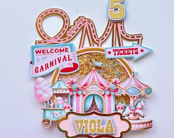 May include: A pink and gold carnival-themed cake topper with a roller coaster, a circus tent, a cotton candy stand, and a carousel. The topper also includes the number 5, the words "Welcome to the Carnival", "Tickets", and the name "Viola".