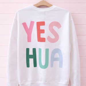 May include: White sweatshirt with the word "YES HUA" printed on the back in pink, red, light pink, green, light green, and light blue block letters. The sweatshirt is hanging on a wooden hanger against a pink background.