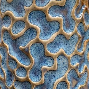 May include: Close-up of a ceramic vase with a textured surface. The design features a network of tan-colored, organic shapes against a speckled blue background. The glaze has a glossy finish, enhancing the visual depth and detail.