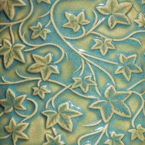 Könnte beinhalten: Nahaufnahme einer Keramikoberfläche mit einem sich wiederholenden Muster aus Efeublättern und -ranken. Die Blätter sind hellgrün, der Hintergrund ist türkis und hellgrün. Die Ranken sind cremeweiß.