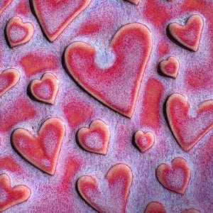 May include: Close-up of a textured surface with raised heart shapes. The hearts vary in size and are primarily red with a lighter, textured appearance. The background is a blend of pink and purple.