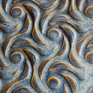 May include: Close-up of a ceramic tile with a repeating pattern. The design features swirling, leaf-like shapes in a bronze color against a mottled blue and white background. Circular accents are interspersed throughout the design, creating a textured surface.