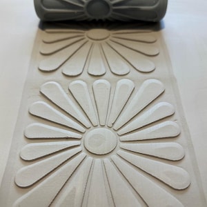 Puede incluir: Un rodillo de pintura gris con un patrón de margarita, junto al patrón en relieve que crea. El rodillo tiene un mango acanalado y un diseño floral. El patrón muestra flores en forma de margarita.