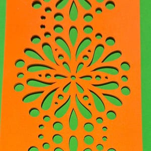 Peut inclure: Gros plan sur un panneau décoratif orange avec un motif floral découpé. Le panneau est posé sur un fond vert. Le motif découpé comprend des formes en goutte et circulaires, créant un design symétrique.