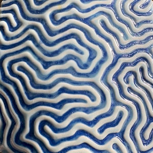 May include: A close-up of a ceramic tile with a blue and white pattern. The design features a series of curved, wave-like lines, creating a textured, organic appearance. The glossy finish enhances the contrast between the colours.