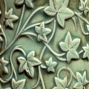 May include: Close-up of a ceramic surface with a repeating ivy leaf pattern. The leaves are light green with darker green shading, and the vines are a lighter colour. The background is a mottled green.