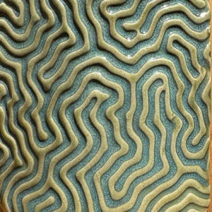 May include: Close-up of a ceramic tile with a textured, organic design. The tile features a pattern of raised, winding lines in a light olive green color, set against a teal blue background. The overall effect is reminiscent of a topographical map.