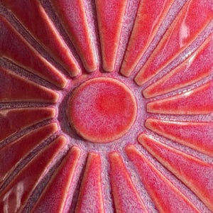 Puede incluir: Primer plano de una pieza de cerámica con un diseño de sol. El centro es un círculo sólido y texturizado, rodeado de formas triangulares radiantes. La paleta de colores es una mezcla de tonos rojo intenso y rosa, con un acabado brillante.