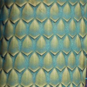 May include: A ceramic surface featuring a repeating three-dimensional scale pattern. The texture consists of raised, pointed, petal-like shapes in a pale yellow-green hue, contrasted by deep teal blue glaze filling the recessed areas between.
