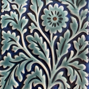 May include: Square ceramic tile with a floral pattern in shades of blue and teal. The design features stylized leaves and a central flower with a brown centre. The tile has a glossy finish.