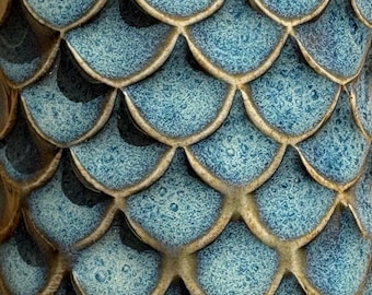 May include: Close-up of a textured, ceramic surface with a repeating pattern of overlapping, teal-colored scales. The scales have a speckled appearance and are outlined with a light brown glaze, creating a dimensional effect.