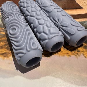 May include: Three cylindrical grey paint rollers featuring distinct embossed patterns. The left roller has a swirling circular design, the middle roller displays a textured pebble-like pattern, and the right roller features a floral motif with petals.