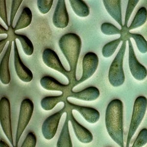 May include: Close-up of a ceramic surface with a repeating floral pattern. The design features stylized flowers and teardrop shapes in varying shades of green and teal. The raised elements create a textured, tactile appearance.