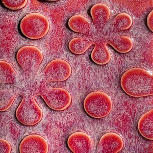 May include: Close-up of a textured surface with a pattern of raised, red, circular and petal-shaped designs. The background is a mottled, light purple and white. The raised elements have a glossy finish.