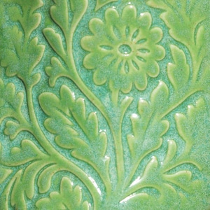 May include: Close-up of a green ceramic tile with a floral relief pattern. The design features a central flower with surrounding leaves and stems, all in a raised, textured finish. The background is a lighter shade of green, creating a contrast with the raised elements.