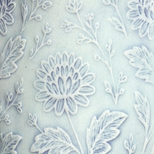 May include: Close-up of a pale blue ceramic surface with a raised floral pattern. The design features large stylized flowers, leaves, and delicate sprigs, creating a textured, embossed effect. The overall aesthetic is reminiscent of vintage or antique decor.