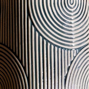 May include: Close-up of a ceramic surface with a repeating pattern of curved lines and vertical stripes. The design features alternating bands of off-white and dark teal, creating a textured, three-dimensional effect. The overall aesthetic is modern and geometric.