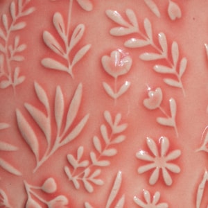 May include: Close-up of a pink surface with a repeating floral pattern. The design features raised white botanical elements, including leaves, flowers, and sprigs. The overall aesthetic is delicate and decorative, with a focus on texture and detail.