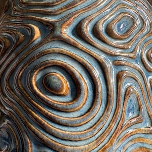 May include: Close-up of a ceramic object with a textured surface. The design features concentric, organic shapes in shades of blue, brown, and gold. The glossy finish enhances the depth and visual interest of the piece.