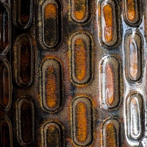May include: Close-up of a textured ceramic surface with a repeating pattern of elongated ovals. The ovals are outlined in dark brown and filled with shades of orange and brown, creating a rustic, earthy aesthetic.